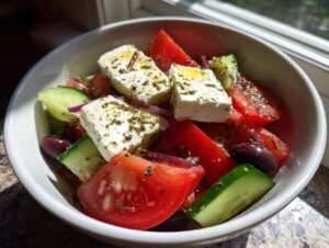 A close-up view of a fresh greek salad featuring large chunks of tomato, cucumber, Kalamata olives, and thick slices of feta cheese drizzled with oil and herbs.