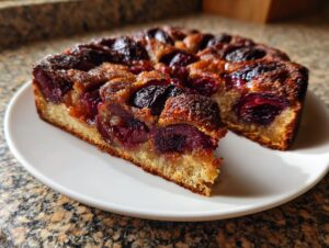 A close-up of a slice of amazing german plum cake, featuring dark baked plums on a golden crust, served on a white plate.