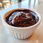 A close-up of rich, glossy chocolate ganache swirled inside a small white ramekin.