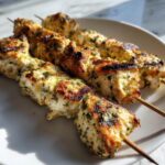 Close-up of three grilled garlic parmesan chicken skewers on a white plate, showing char marks and herbs.
