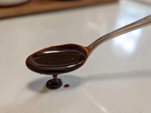 Close-up of thick, glossy homemade chocolate syrup dripping from a silver spoon onto a light countertop.