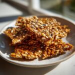 A stack of glossy, golden-brown pieces of homemade seed brittle, featuring sunflower and pumpkin seeds, resting on a light ceramic plate.