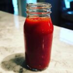 A glass bottle filled with vibrant red homemade hot sauce sitting on a light countertop.