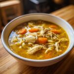 Close-up of a white bowl filled with rich, yellow broth, shredded chicken, egg noodles, and bright orange carrots from the slow cooker chicken noodle soup.