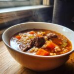 A close-up of a steaming white bowl filled with rich beef barley soup, featuring chunks of beef, carrots, and barley.
