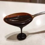 Close-up of thick, glossy chocolate syrup dripping from a silver spoon onto a light countertop.