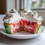 A close-up, cross-section view of a watermelon cupcake revealing the pink interior, white frosting, and black sprinkles, resembling a watermelon slice.