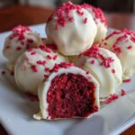 A close-up of white chocolate-dipped red velvet cake balls, one cut in half showing the rich red interior.