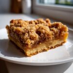 A close-up, sunlit shot of a single, thick square of apple pie bars with a crumbly topping on a white plate.