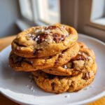 A stack of three thick, golden Bakery style chocolate chip cookies topped with melted chocolate and flaky sea salt.