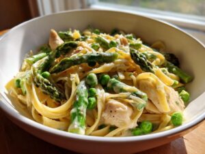 A close-up of creamy Chicken primavera pasta featuring fettuccine, chunks of chicken, bright green asparagus spears, and peas.