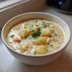 A close-up of a creamy white bowl filled with thick clam chowder, featuring chunks of potato and carrots, topped with fresh parsley.