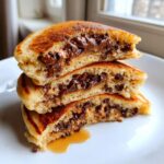 A stack of three fluffy chocolate chip pancakes cut in half, revealing melted chocolate centers and syrup pooling below.