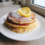 A stack of three fluffy Lemon ricotta pancakes dusted heavily with powdered sugar and topped with a fresh lemon slice.