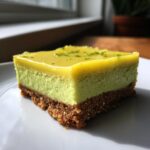 A close-up of a single, thick square of Key lime pie bars showing a graham cracker crust and bright green filling topped with glaze and lime zest.