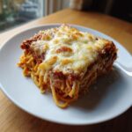 A square slice of baked leftover spaghetti bake topped with melted, golden cheese on a white plate.