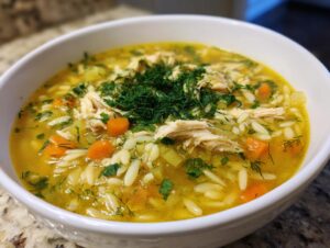 Close-up of a white bowl filled with Mediterranean lemon chicken soup featuring shredded chicken, orzo pasta, carrots, and fresh dill.