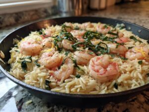 Close-up of creamy one pan shrimp orzo topped with pink shrimp, fresh basil, and lemon zest in a dark skillet.