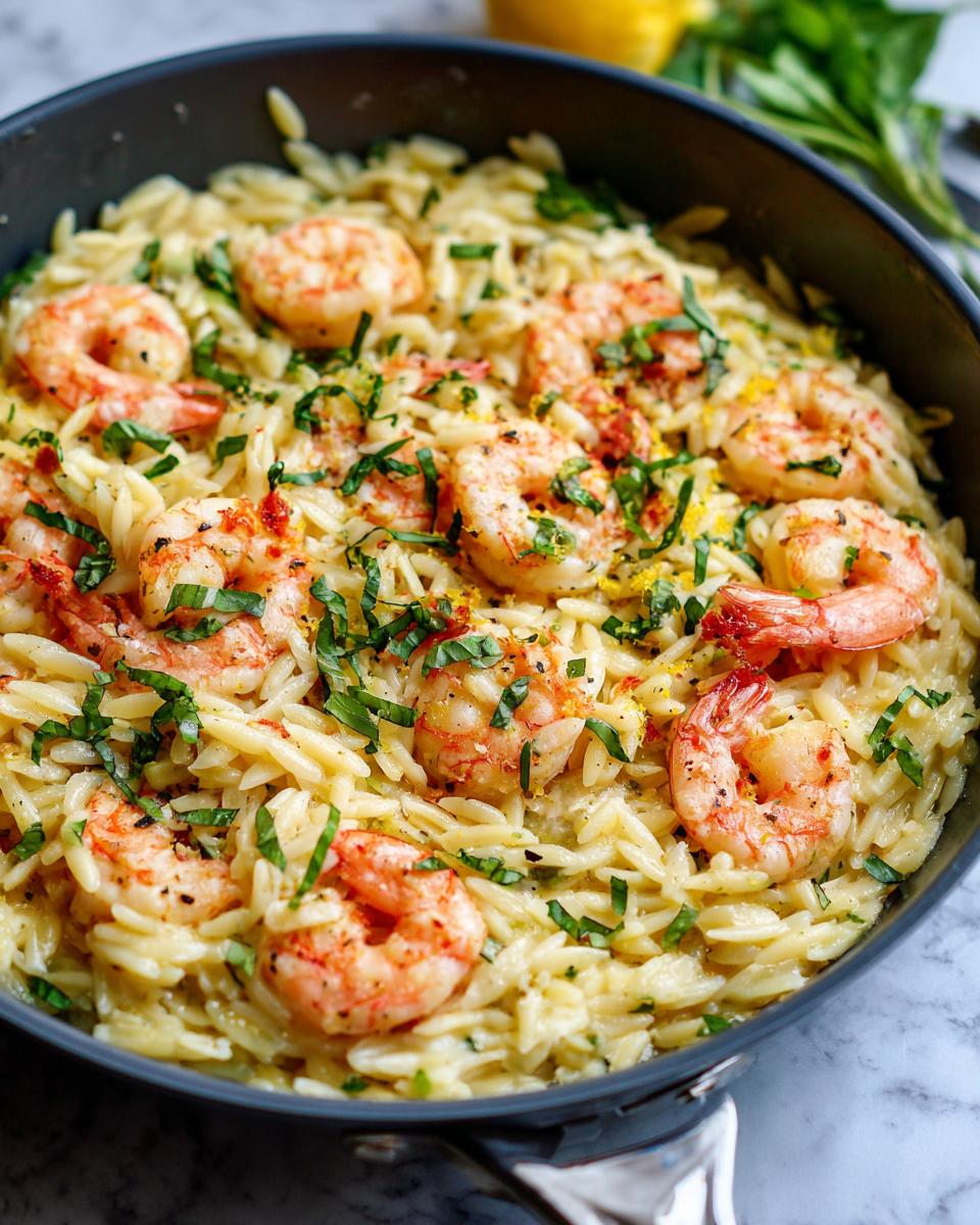 Close-up of creamy one pan shrimp orzo garnished with fresh basil and lemon zest in a dark skillet.