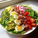 A beautifully arranged Spring Cobb salad featuring hard-boiled eggs, avocado, bacon, radishes, tomatoes, and snap peas on lettuce.