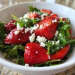 Close-up of a fresh strawberry arugula salad topped with crumbled white feta cheese in a white bowl.